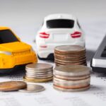 Two toy cars sit on a sheet of paper featuring many numbers. They are next to multiple stacks of coins and a calculator.