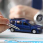 Close-up of a man's hand holding a silver pen over a form on a clipboard. The form has a toy car on top.