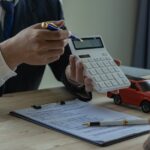 A man wearing a business suit behind a desk holds up a calculator. A toy car and a clipboard with a contract lie on the desk.