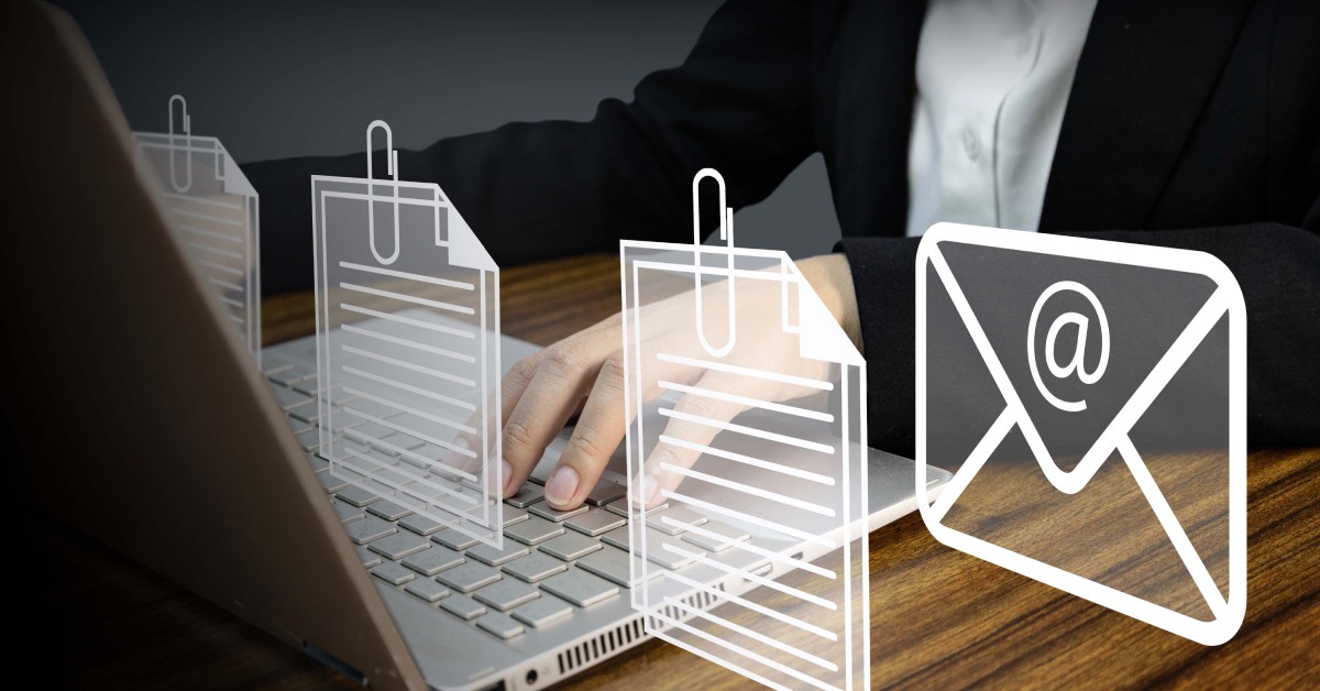 A person works on their laptop on a desk. Symbols of an email and attached documents float above the laptop.