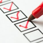 Close-up of the end of a red pen, as someone uses it to make checks in small boxes on a white piece of paper.