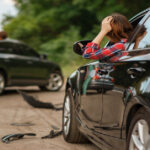 A woman sits behind the wheel of a car after an accident and holds her head. A man examines the damage to his car.