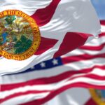 A Florida state flag waves in the air on a flagpole. It stands next to a second flagpole, featuring the U.S. flag.