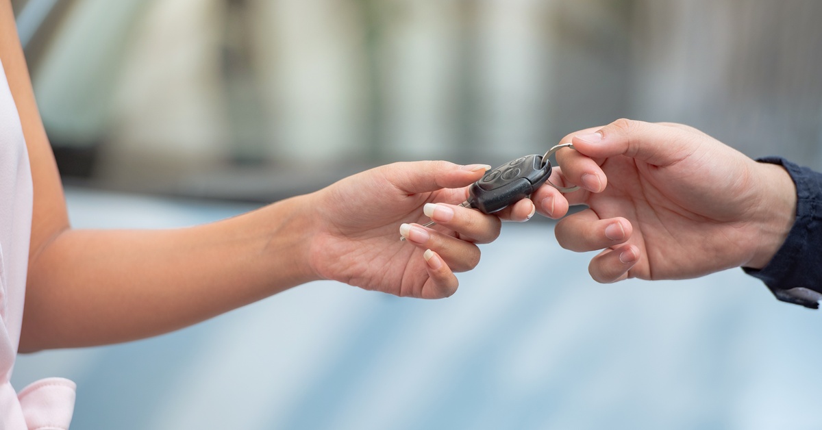 Close-up of a person's hand extending car keys to another. One person wears blue, while the other wears pink.
