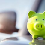 A small green piggy bank sits on the hood of a parked car. The car has a reddish color and sits in front of a white wall.