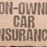 A hand holds up a sign on a piece of cardboard in front of a building. The sign reads "Non-owner car insurance."