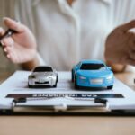 Two toy cars sit on top of documents sitting on a clipboard on a desk. The document reads "car insurance."