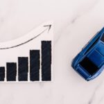 A blue toy car sits on a marble surface next to a bar graph. Both the bars and an arrow above them are trending upward.