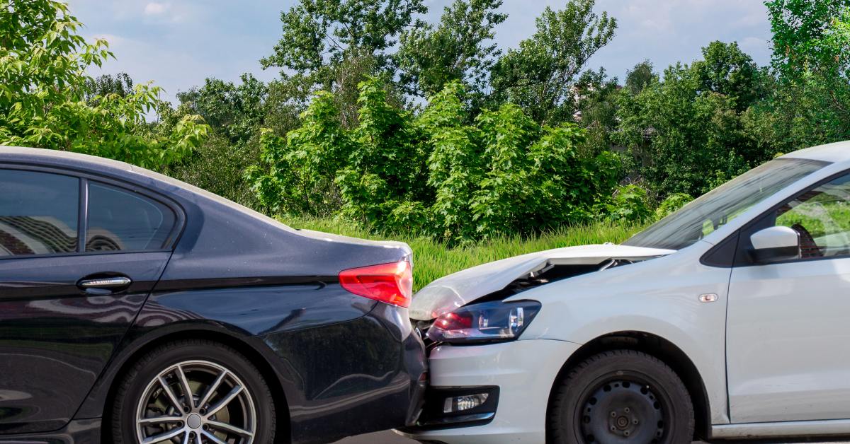 A white car has crashed into the back of a black car. Several trees stand in the background behind the wreck.