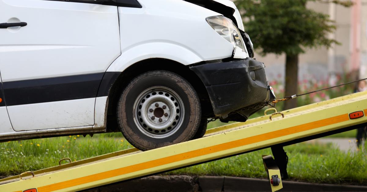 A metal wire pulls a wrecked white van up a yellow ramp. A tree and various plants sit in the background.