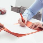 Close-up of a woman's hands as she signs a document on a clipboard. A toy car sits on the table nearby.