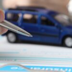 A hand holds a pen over a car insurance form on a clipboard. A blue toy car also lies on the clipboard.