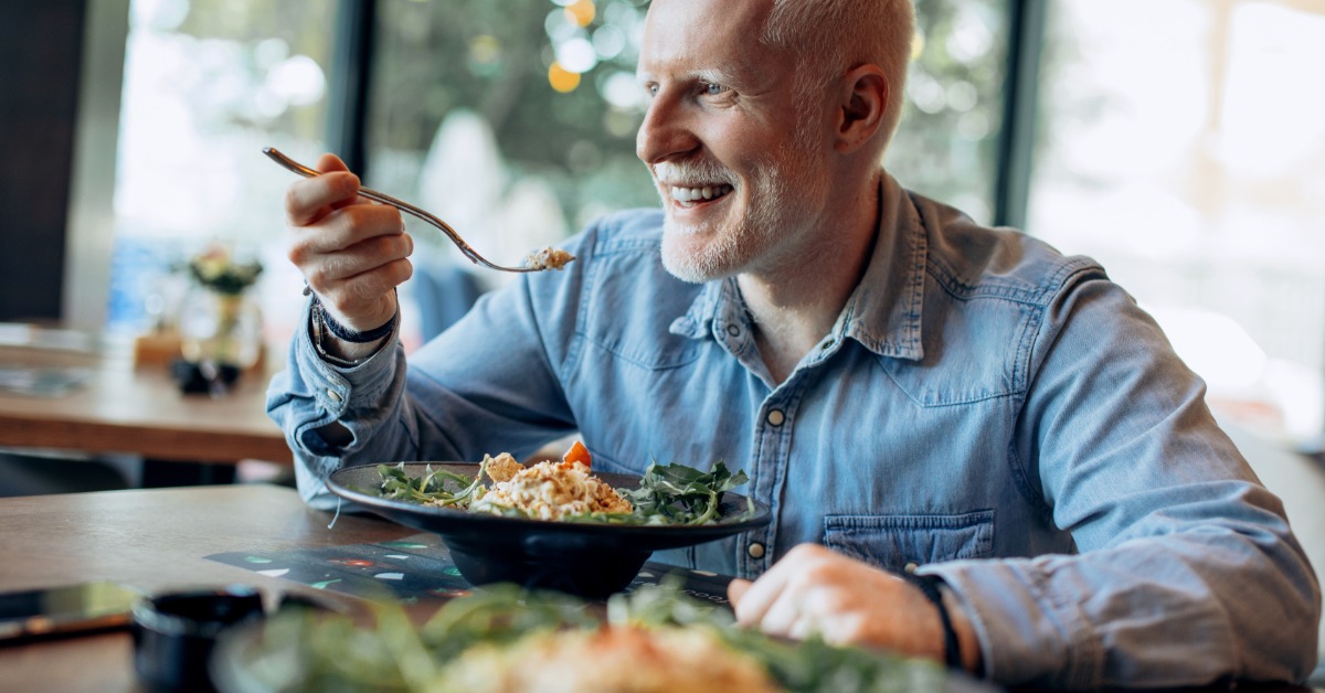 A man smiles as he prepares to enjoy a salad while sitting in a restaurant. He holds a fork and wears a denim shirt.