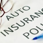 A pen and a pair of glasses lie on a piece of paper that reads the word "Auto Insurance Policy" in big letters.