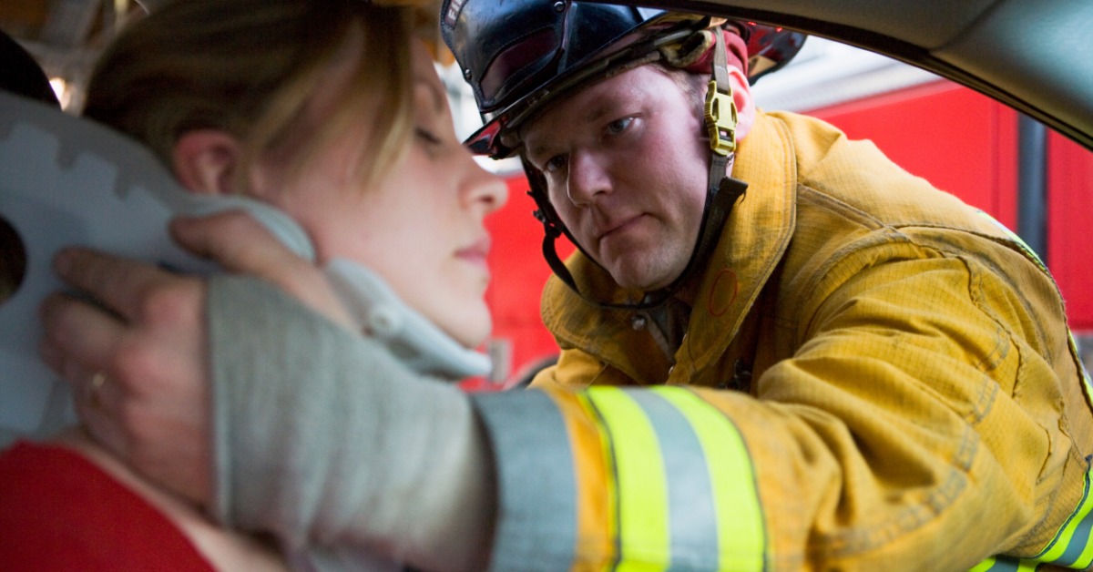 A firefighter helps put a neck brace on a woman who sits behind the wheel of her car. She appears to be in pain.
