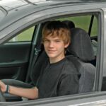 A teenage boy smiles as he sits behind the wheel of an SUV. He wears a black shirt and has his hand on the wheel.