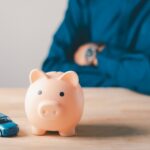 A toy piggy bank and a toy car sit on a wooden table top. A person sits behind them, arms folded and wearing a blue shirt.