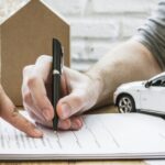 A close-up of a person's hand signing insurance paperwork on a desk. Another hand points where to sign.