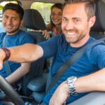 A group of people sits together in a car and smiles. Two people in the front of the vehicle wear blue shirts.