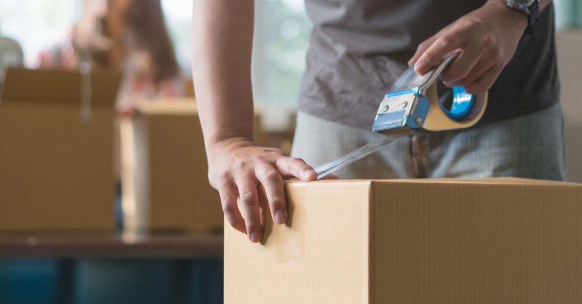 A person seals a cardboard box with clear tape while another person sits near moving boxes in the background.