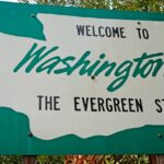 A green Washington welcome sign with white state outline and lettering standing on wooden posts beside leafy trees.