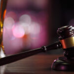 A tall glass of beer sits next to a gavel and car key resting on a wooden sound block on a wood countertop.