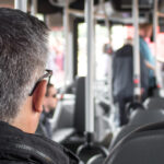 A man with grey hair and glasses sits on a public bus. In the blurry background, two people are leaving the bus.