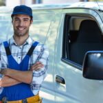 A male carpenter in a uniform and toolbelt stands with crossed arms, smiling in front of a white work van.