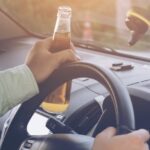 A man wearing a long-sleeved shirt holds a beer bottle in his left hand while steering a car with his right hand.