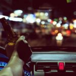 A man driving a car while holding a beer bottle in his right hand stopped in traffic at night on a busy street.