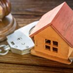 A small wooden house inside an open metal handcuff next to a wooden gavel and sound block on a wooden table.