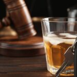 A car key in front of a glass filled with an alcoholic beverage with a gavel and gold scale in the blurred background.