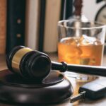 A gavel resting on a sound block near a car key and a glass of an alcoholic drink on a wooden desk with books.