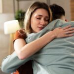 A woman is embracing a man with her head on his shoulder while both are sitting on a couch in a living room.