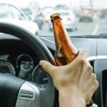 A person driving a car in traffic. They are holding an open beer bottle in their right hand, which rests on the steering wheel.