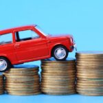 A red toy car on top of four ascending stacks of coins from left to right on a solid light blue background.