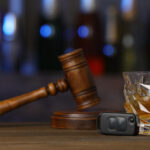 A wooden gavel rests on a sound block next to a car key and an alcoholic drink on a wood table, with a blurred background.