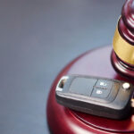 A close-up of a gavel resting on a wooden sound block next to a black car key on a dark hardwood desk.