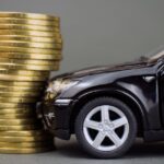 A miniature black model car colliding with a tall stack of gleaming gold coins, set against a gray background.