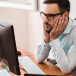 A man works at a desktop computer in an office. He faces a monitor, looking confused, his hands on his chin.