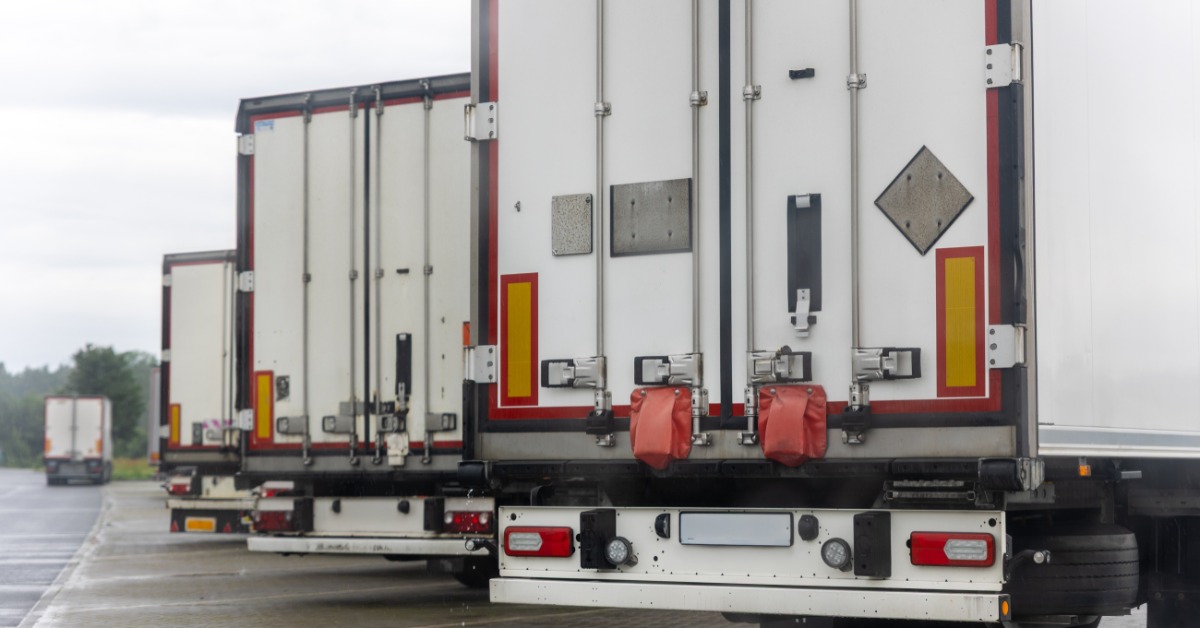 A row of white truck trailers parked on wet asphalt. One of the trucks drives away from the other vehicles.