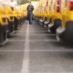 A person in a blue jumpsuit walks between repeated rows of yellow vans parked outdoors near a building.