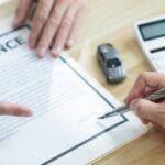 A man points to a clipboard holding a document that says "insurance" while another person prepares to sign it.