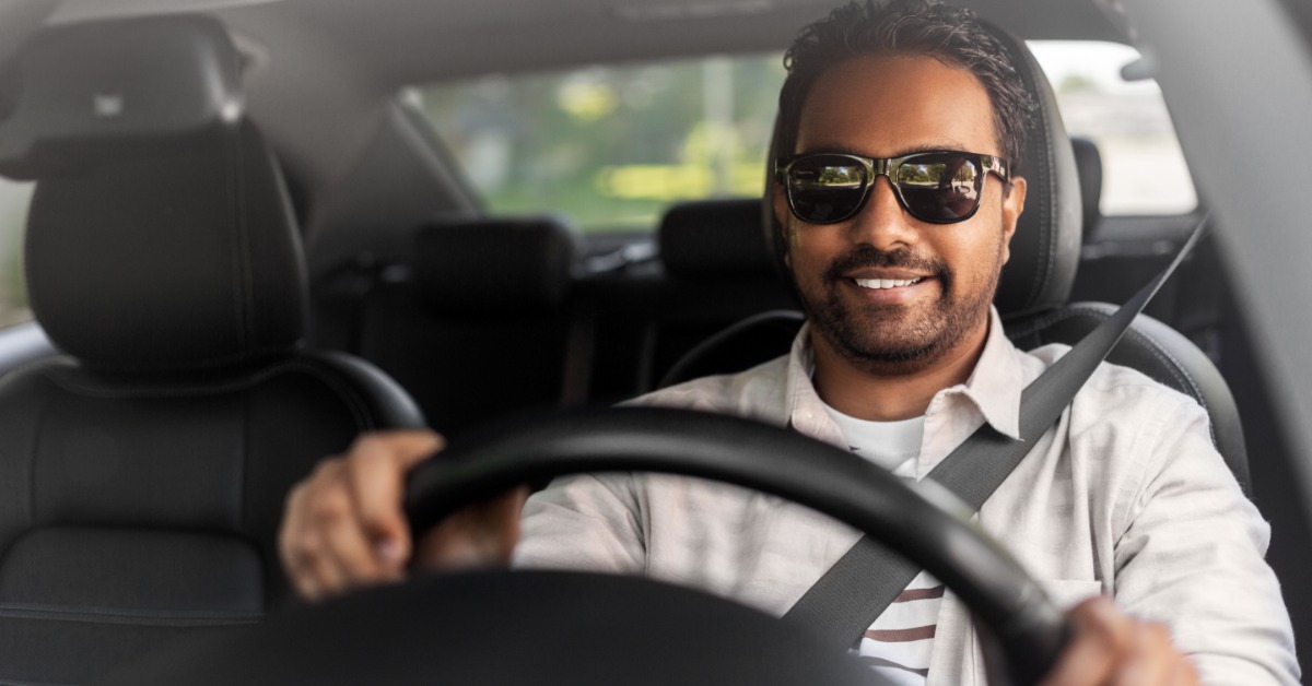 A man wears sunglasses and sits in the driver's seat of a car. He has both hands on the steering wheel.