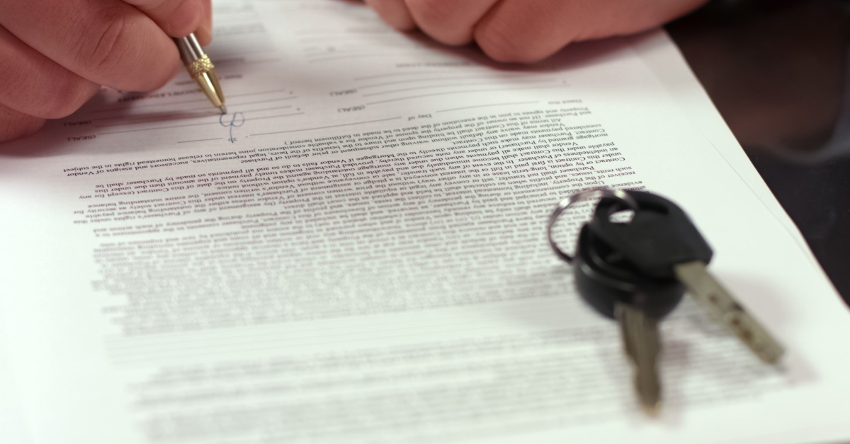 Close-up of a person signing paperwork with a pen on a table. A set of car keys lies on top of the documents.