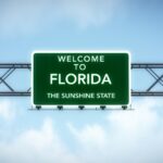 A green "Welcome to Florida" highway sign hangs over a metal gantry with blue sky and clouds behind it.