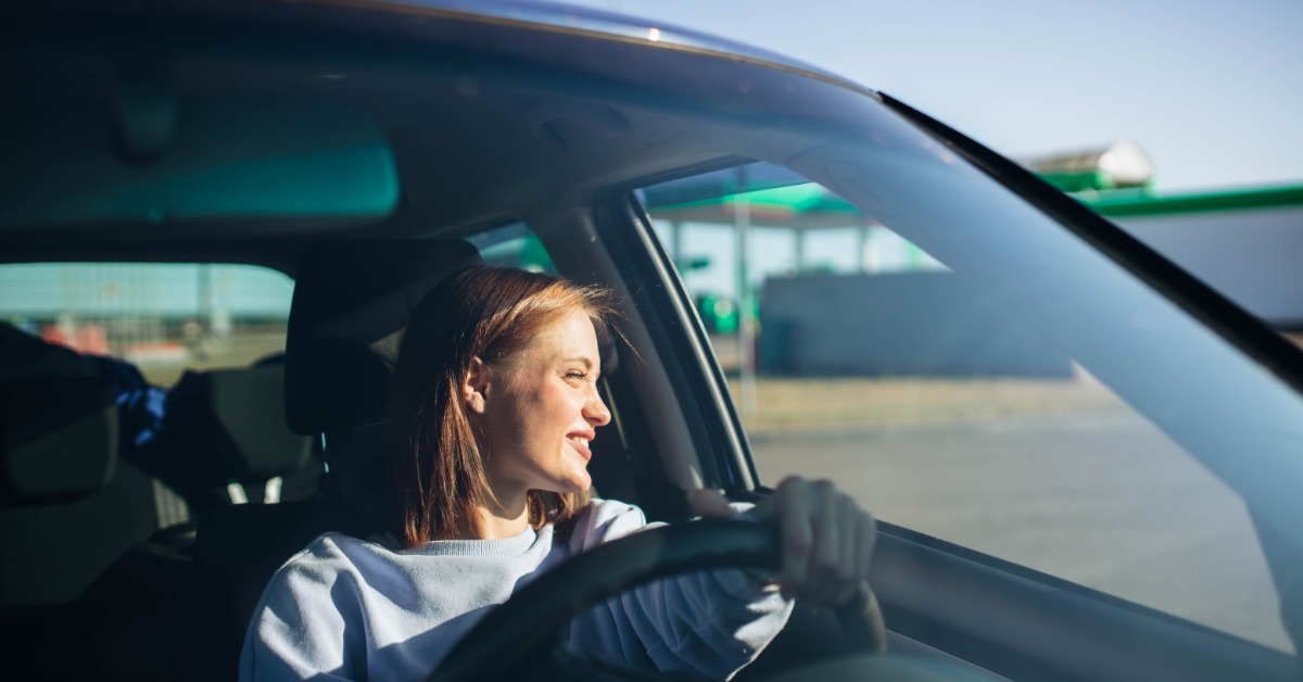 A woman smiles while driving a car. She holds the steering wheel as sunlight enters through the windshield.