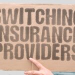 A hand holds a cardboard sign that reads "switching insurance providers" in front of blurred office buildings.