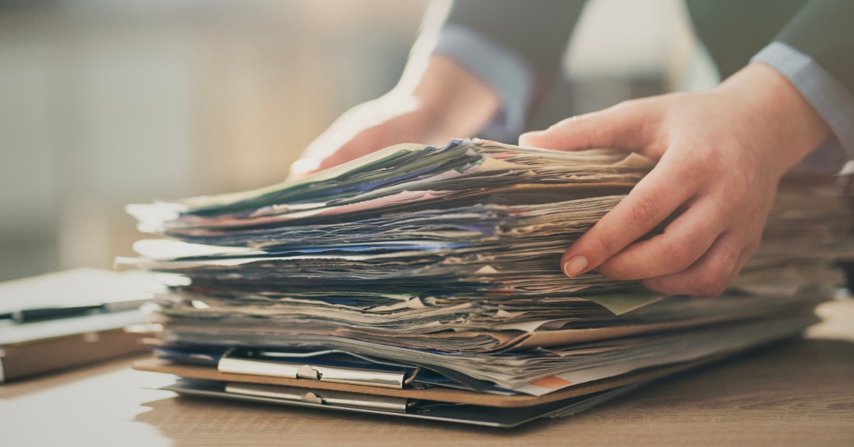 Close-up of a person's hands setting a large pile of file folders onto a wooden desk. The person wears a sports coat.