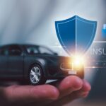 A hand holding a small black toy car. The word "insurance" appears next to it with a blue shield and folders.